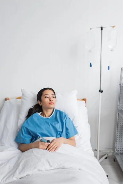 Thoughtful asian woman holding inhaler and looking away on bed in hospital ward — Stock Photo