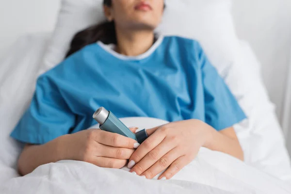 Vista recortada de la mujer borrosa sosteniendo el inhalador mientras está acostada en la cama en la clínica - foto de stock