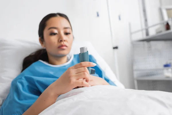 Pensive asian woman looking at inhaler while lying on bed in hospital — Stock Photo