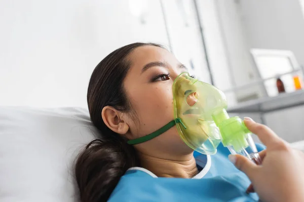 Young asian woman in oxygen mask lying on hospital bed and looking away — Stock Photo
