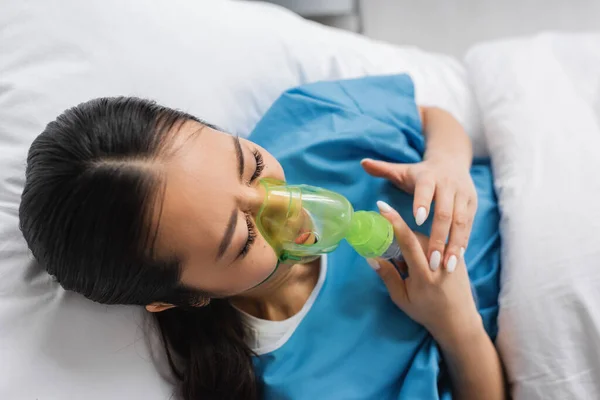 Top view of ill asian woman in oxygen mask lying on bed in hospital ward — Stock Photo