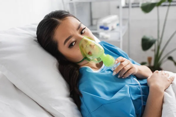 Junge Asiatin schaut weg, während sie in Sauerstoffmaske auf dem Krankenhausbett liegt — Stockfoto