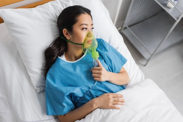 Vista dall'alto di sconvolto asiatico donna sdraiata su ospedale letto in ossigeno maschera e guardando altrove — Foto stock
