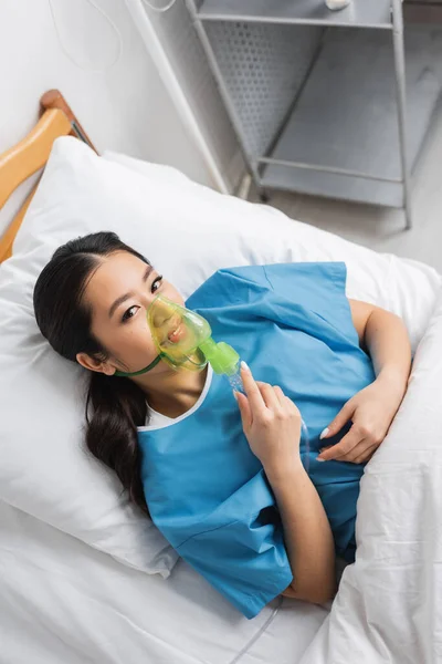 Вид сверху положительной азиатской женщины, дышащей кислородной маской и смотрящей на камеру на кровати в клинике — стоковое фото