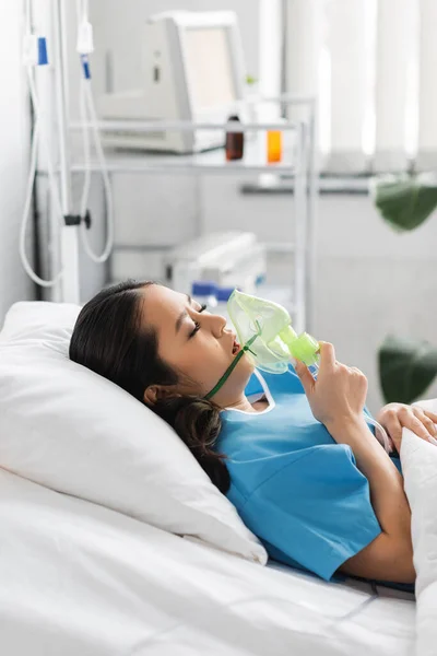 Side view of sick asian woman holding oxygen mask while lying on bed in hospital ward — Stock Photo