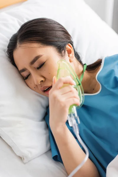 Doente asiático mulher segurando oxigênio máscara enquanto deitado no hospital cama com olhos fechados — Fotografia de Stock