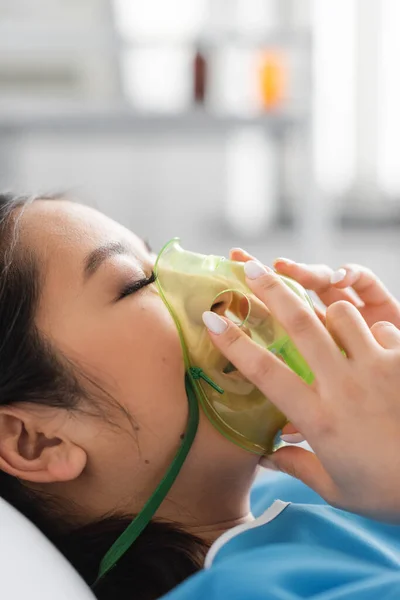 Vista lateral de la mujer asiática enferma tocando la máscara de oxígeno mientras está acostado en el hospital — Stock Photo