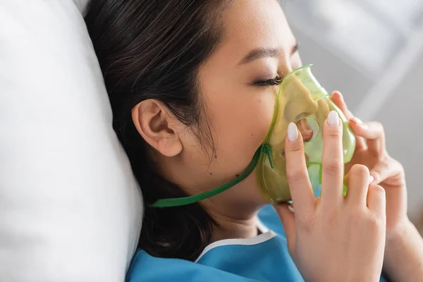 Side view of diseased asian woman adjusting oxygen mask in hospital ward — Stock Photo