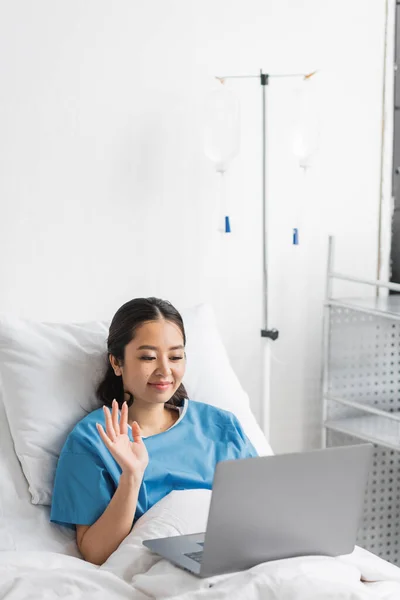 Alegre asiático mujer agitando la mano durante el video chat en portátil en hospital sala - foto de stock