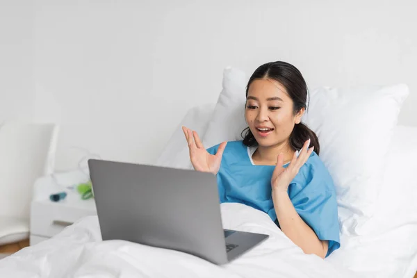 Asombrado asiático mujer mostrando wow gesto durante la videollamada en portátil en hospital cama — Stock Photo