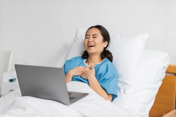 Excitado asiático mujer tocando pecho y riendo con cerrado ojos mientras viendo comedia película en portátil en clínica — Stock Photo