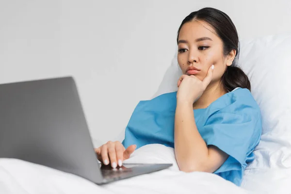 Réfléchie asiatique femme couché sur lit d'hôpital et regardant ordinateur portable — Photo de stock