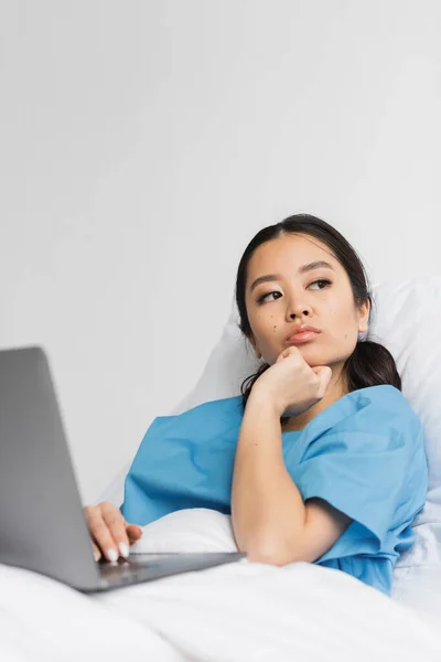 Mujer asiática pensativa sentada en la cama del hospital con portátil y mirando hacia otro lado - foto de stock