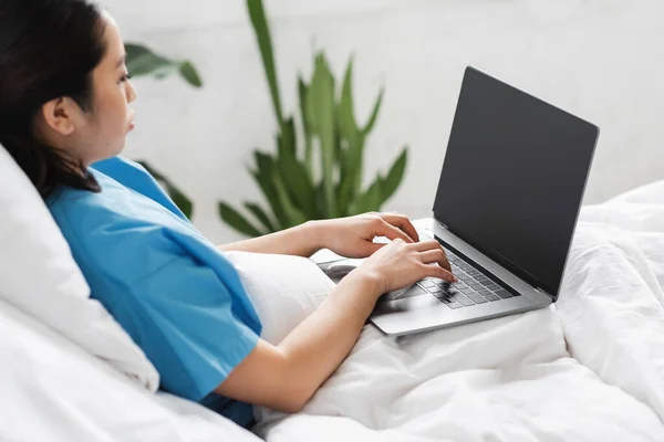 Young asian woman sitting on hospital bed and typing on laptop with blank screen — Stock Photo