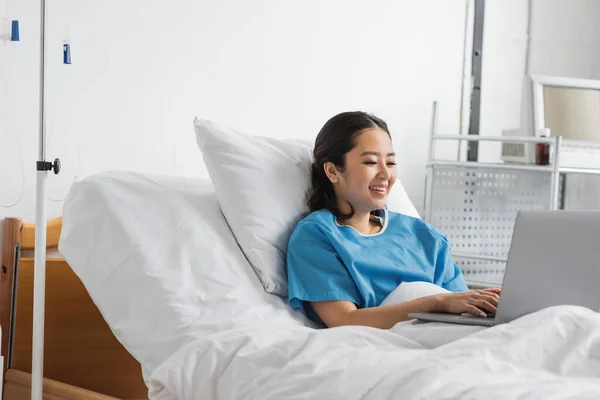 Freudige Asiatin tippt im Krankenhausbett auf Laptop — Stockfoto