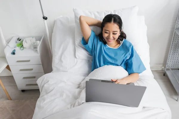 Alto ángulo vista de alegre asiático mujer usando laptop en cama en clínica - foto de stock