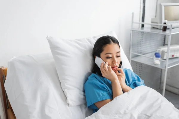 Mujer asiática molesta sosteniendo la mano cerca de la cara mientras habla en el teléfono móvil en la sala de hospital - foto de stock