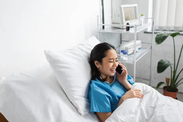 Alegre asiático mujer sonriendo con cerrado ojos mientras hablando en celular en hospital sala - foto de stock