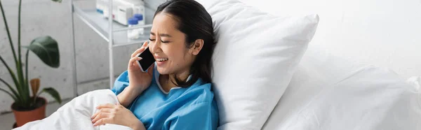 Радісна азіатська жінка розмовляє на мобільному телефоні і посміхається з закритими очима на ліжку в клініці, банер — стокове фото