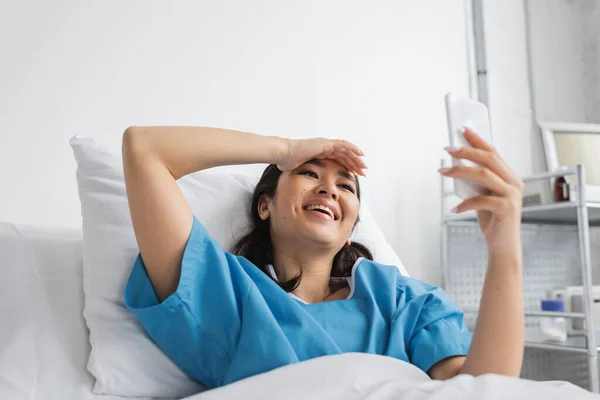 Excitada mujer asiática tocando la frente y mirando el teléfono inteligente en la cama de hospital - foto de stock