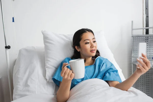Surprised asian woman with cup of tea looking at mobile phone in hospital ward — Stock Photo