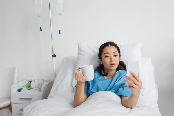 Mujer asiática sorprendida sosteniendo taza de té y mirando el teléfono móvil en la cama en la clínica - foto de stock