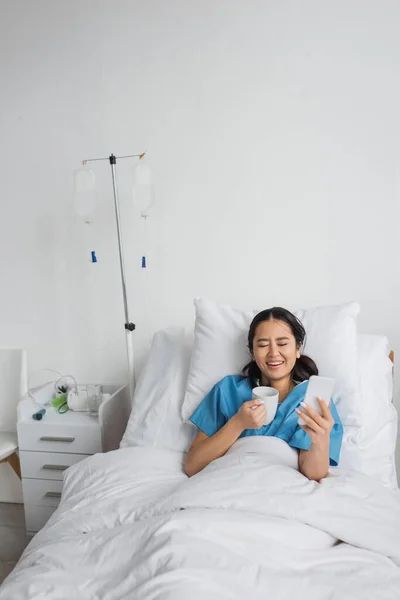 Excited asian woman with smartphone and cup of tea laughing with closed eyes on hospital bed — Stock Photo