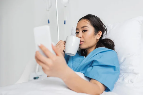 Young asian woman drinking tea and looking at blurred cellphone in hospital ward — Stock Photo