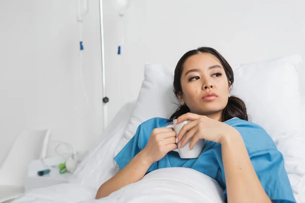 Pensativo asiático mujer holding té taza y mirando lejos en hospital cama - foto de stock