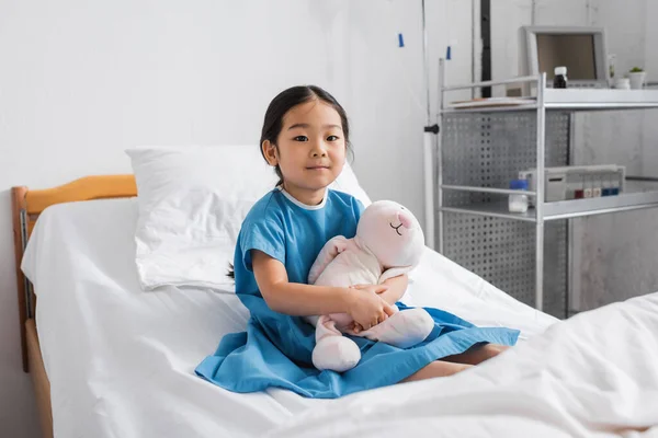 Позитивна азіатська дівчина обіймає іграшкового кролика і дивиться на камеру, сидячи на лікарняному ліжку — стокове фото