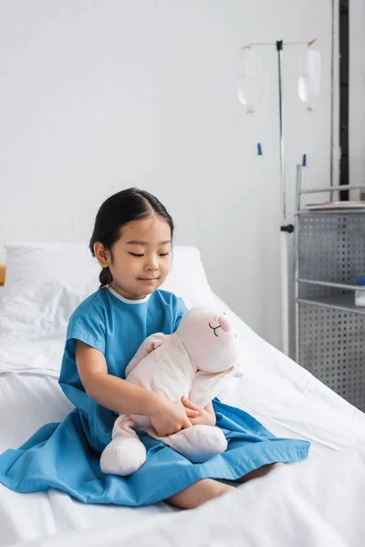 Smiling asian girl sitting on bed in hospital ward and hugging toy bunny — Stock Photo