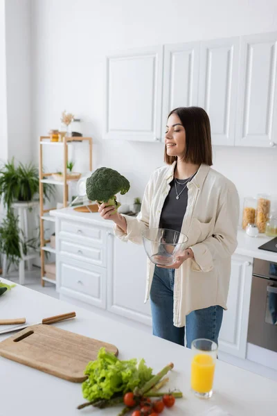 Улыбающаяся женщина с брокколи и миской рядом с овощами и разделочной доской на кухне — стоковое фото