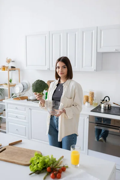 Усміхнена жінка тримає брокколі і миску, дивлячись на камеру на кухні — стокове фото
