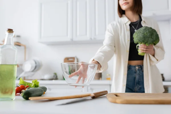 Cropped view of blurred woman holding bowl and broccoli in kitchen — Stock Photo