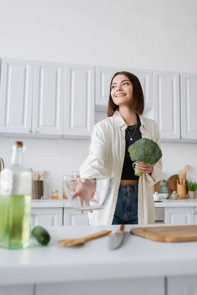 Mulher positiva segurando brócolis e tigela borrada na cozinha — Fotografia de Stock