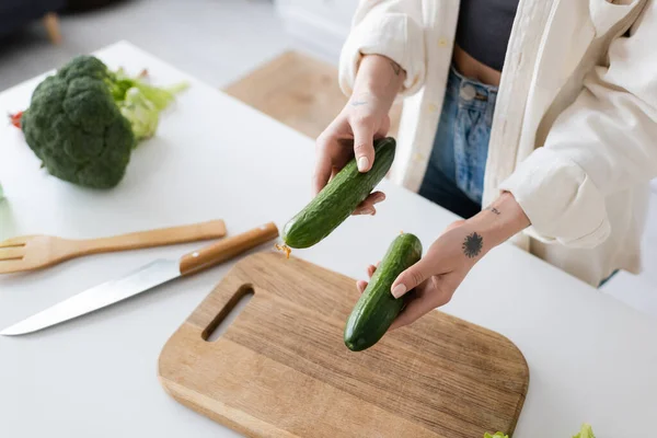 Cropped view of tattooed woman holding cucumbers near cutting board in kitchen — Stock Photo