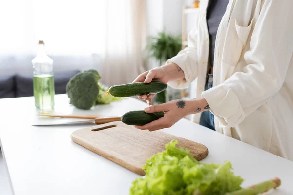 Cropped view of tattooed woman holding cucumbers near vegetables and cutting board in kitchen — Stock Photo
