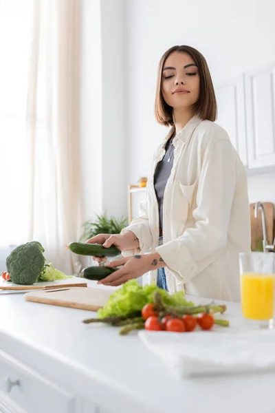Брюнетка держит огурцы возле размытых овощей и апельсиновый сок на кухне — стоковое фото