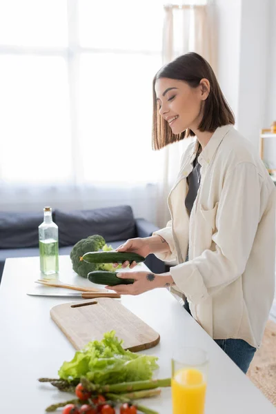 Smiling woman holding cucumbers near vegetables and cutting board in kitchen — Stock Photo