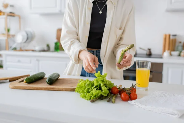 Cropped view of woman holding asparagus near vegetables and orange juice in kitchen — Stock Photo