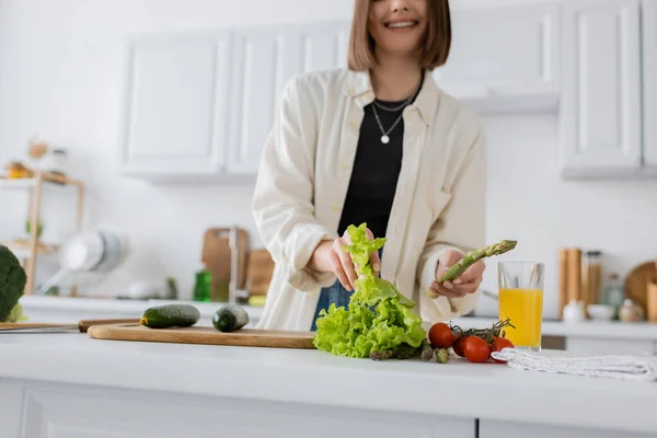Cropped view of blurred smiling woman holding asparagus and lettuce in kitchen — Stock Photo