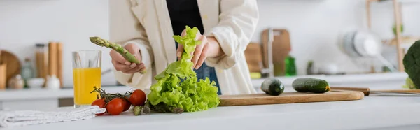 Обрезанный вид молодой женщины, держащей спаржу и салат на кухне, баннер — стоковое фото
