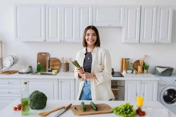 Mulher alegre segurando espargos e olhando para a câmera perto de legumes na cozinha — Fotografia de Stock