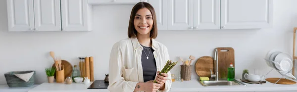 Carefree woman looking at camera and holding asparagus in kitchen, banner — Stock Photo