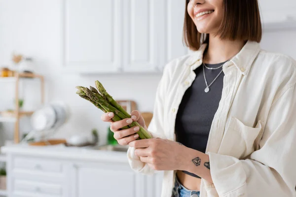 Vista recortada de mujer alegre tatuada sosteniendo espárragos en la cocina - foto de stock