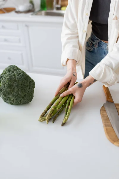 Cropped view of tattooed woman holding asparagus near broccoli and cutting board in kitchen — Stock Photo