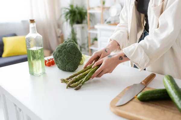 Vista ritagliata della donna tatuata che mette gli asparagi sul tavolo vicino all'olio d'oliva e al tagliere in cucina — Foto stock