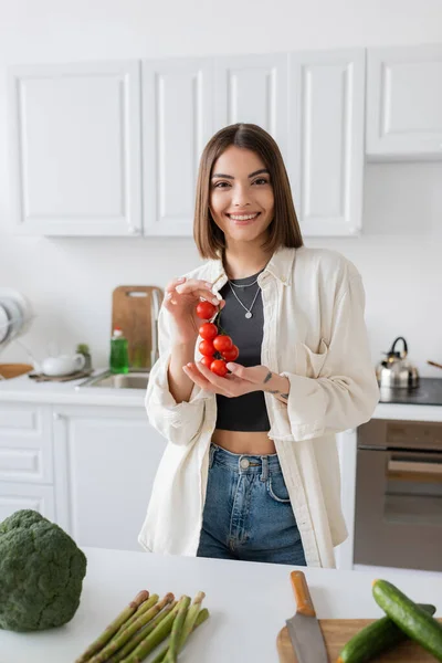Smiling young woman holding cherry tomatoes near vegetables in kitchen — Stock Photo