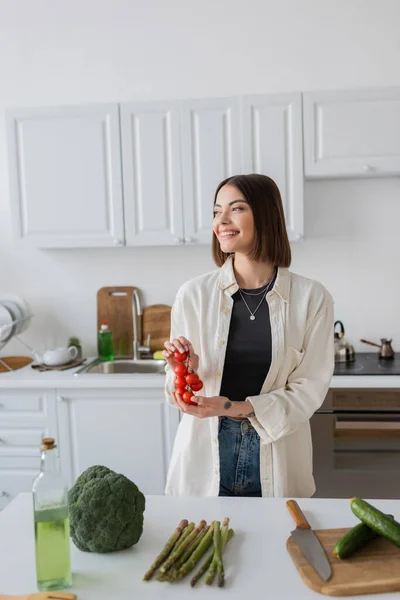 Glückliche junge Frau hält Kirschtomaten neben frischem Gemüse und Olivenöl in der Küche — Stockfoto