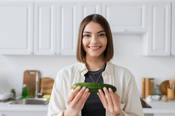Lächelnde brünette Frau mit Gurke in der Hand und Blick in die Kamera in der Küche — Stockfoto